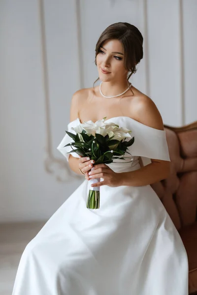ブルネット花嫁でタイトウェディングドレスで明るいスタジオで腕椅子に花束 — ストック写真