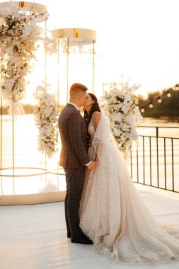 Gelin ve damat, nehrin kıyısındaki bir iskelede sarı bir günbatımının arkaplanına karşı.