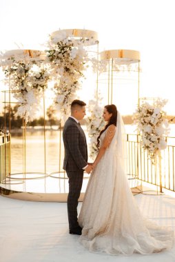 Gelin ve damat, nehrin kıyısındaki bir iskelede sarı bir günbatımının arkaplanına karşı.