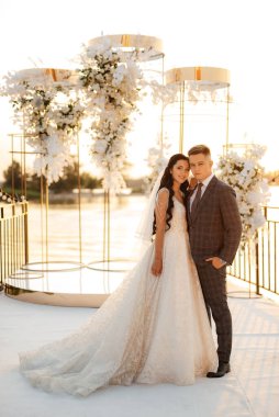 Gelin ve damat, nehrin kıyısındaki bir iskelede sarı bir günbatımının arkaplanına karşı.