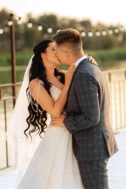 Gelin ve damat, nehrin kıyısındaki bir iskelede sarı bir günbatımının arkaplanına karşı.