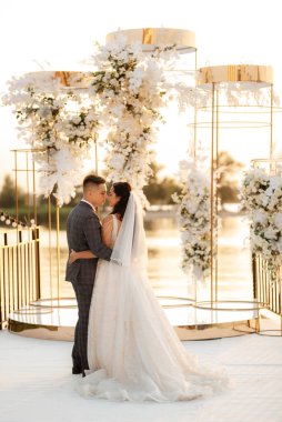 Gelin ve damat, nehrin kıyısındaki bir iskelede sarı bir günbatımının arkaplanına karşı.