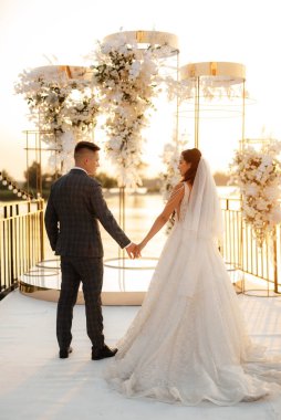 Gelin ve damat, nehrin kıyısındaki bir iskelede sarı bir günbatımının arkaplanına karşı.