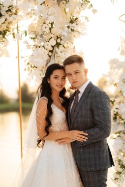 Gelin ve damat, nehrin kıyısındaki bir iskelede sarı bir günbatımının arkaplanına karşı.