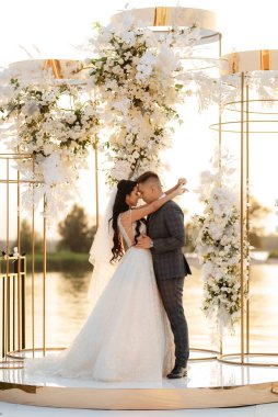 Gelin ve damat, nehrin kıyısındaki bir iskelede sarı bir günbatımının arkaplanına karşı.