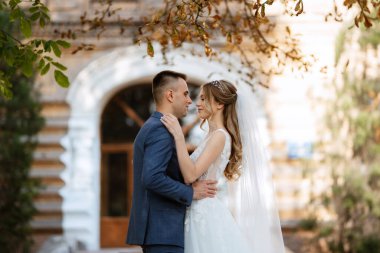 Yeni evliler, eski binaların yakınlarındaki ağaçların ve çam ağaçlarının önünde yürüyorlar.