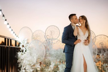 Gelin ve damat, nehrin kıyısındaki bir iskelede sarı bir günbatımının arkaplanına karşı.