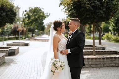 Kahverengi takım elbiseli damat ve şehir ortamında beyaz elbiseli gelin.