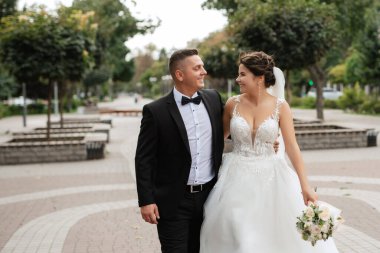 Kahverengi takım elbiseli damat ve şehir ortamında beyaz elbiseli gelin.