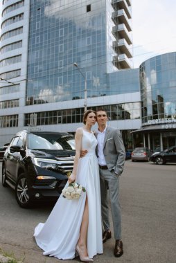 Gelin ve damat cam bir gökdelenin yanında siyah bir arabayla