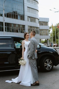 Gelin ve damat cam bir gökdelenin yanında siyah bir arabayla