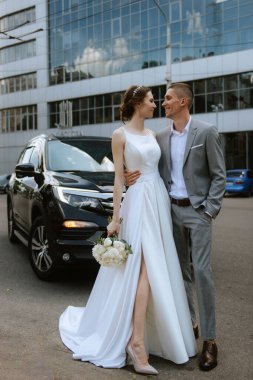 Gelin ve damat cam bir gökdelenin yanında siyah bir arabayla