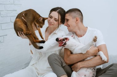 Genç bir çift, parlak bir odada kedi ve köpekle oynuyorlar.