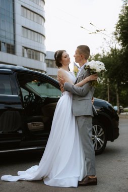 Gelin ve damat cam bir gökdelenin yanında siyah bir arabayla