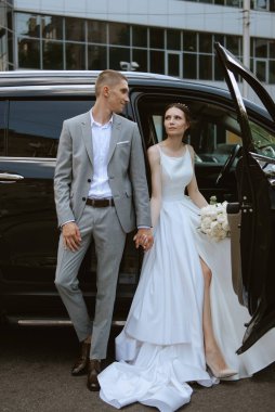 Gelin ve damat cam bir gökdelenin yanında siyah bir arabayla