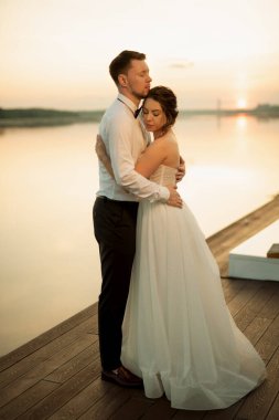 bride and groom against the backdrop of a yellow sunset on a pier near the river