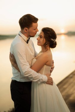 Gelin ve damat, nehrin kıyısındaki bir iskelede sarı bir günbatımının arkaplanına karşı.