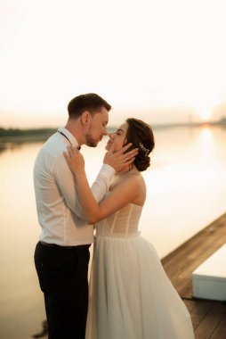 Gelin ve damat, nehrin kıyısındaki bir iskelede sarı bir günbatımının arkaplanına karşı.