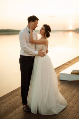 Gelin ve damat, nehrin kıyısındaki bir iskelede sarı bir günbatımının arkaplanına karşı.