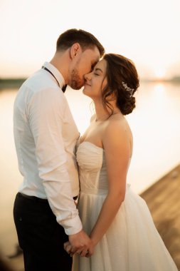 Gelin ve damat, nehrin kıyısındaki bir iskelede sarı bir günbatımının arkaplanına karşı.