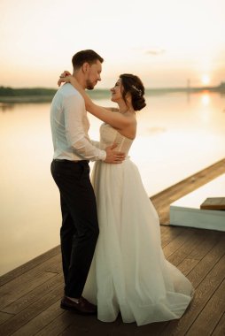 Gelin ve damat, nehrin kıyısındaki bir iskelede sarı bir günbatımının arkaplanına karşı.