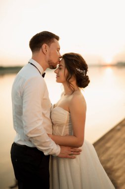 Gelin ve damat, nehrin kıyısındaki bir iskelede sarı bir günbatımının arkaplanına karşı.