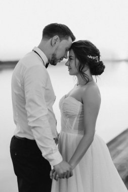 Gelin ve damat, nehrin kıyısındaki bir iskelede sarı bir günbatımının arkaplanına karşı.