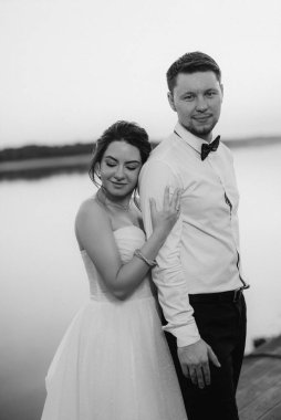 Gelin ve damat, nehrin kıyısındaki bir iskelede sarı bir günbatımının arkaplanına karşı.