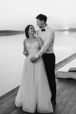 Gelin ve damat, nehrin kıyısındaki bir iskelede sarı bir günbatımının arkaplanına karşı.