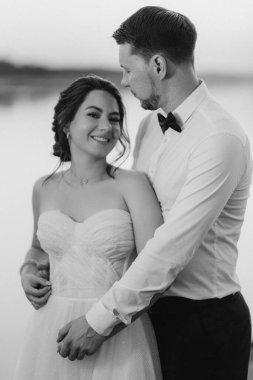 Gelin ve damat, nehrin kıyısındaki bir iskelede sarı bir günbatımının arkaplanına karşı.