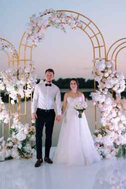 Gelin ve damat, nehrin kıyısındaki bir iskelede sarı bir günbatımının arkaplanına karşı.