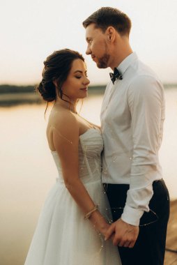 Gelin ve damat, nehrin kıyısındaki bir iskelede sarı bir günbatımının arkaplanına karşı.