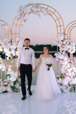 Gelin ve damat, nehrin kıyısındaki bir iskelede sarı bir günbatımının arkaplanına karşı.