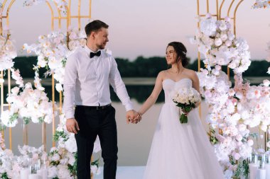 Gelin ve damat, nehrin kıyısındaki bir iskelede sarı bir günbatımının arkaplanına karşı.