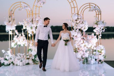 Gelin ve damat, nehrin kıyısındaki bir iskelede sarı bir günbatımının arkaplanına karşı.