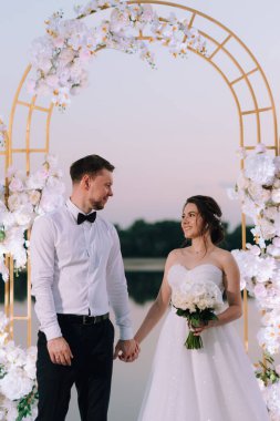 Gelin ve damat, nehrin kıyısındaki bir iskelede sarı bir günbatımının arkaplanına karşı.