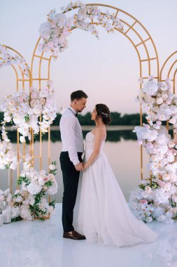 Gelin ve damat, nehrin kıyısındaki bir iskelede sarı bir günbatımının arkaplanına karşı.