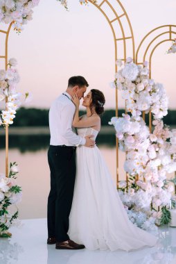 Gelin ve damat, nehrin kıyısındaki bir iskelede sarı bir günbatımının arkaplanına karşı.