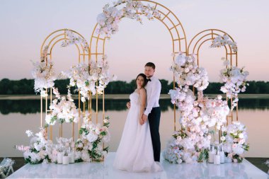 Gelin ve damat, nehrin kıyısındaki bir iskelede sarı bir günbatımının arkaplanına karşı.