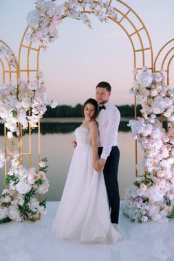 Gelin ve damat, nehrin kıyısındaki bir iskelede sarı bir günbatımının arkaplanına karşı.