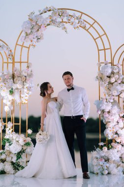 Gelin ve damat, nehrin kıyısındaki bir iskelede sarı bir günbatımının arkaplanına karşı.