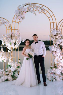 Gelin ve damat, nehrin kıyısındaki bir iskelede sarı bir günbatımının arkaplanına karşı.