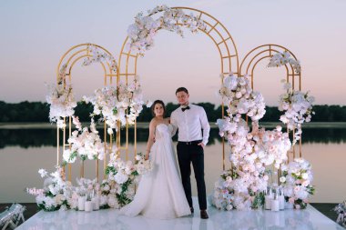 Gelin ve damat, nehrin kıyısındaki bir iskelede sarı bir günbatımının arkaplanına karşı.