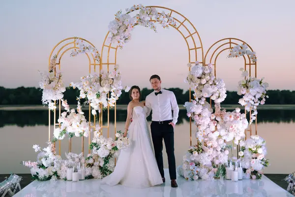 Gelin ve damat, nehrin kıyısındaki bir iskelede sarı bir günbatımının arkaplanına karşı.