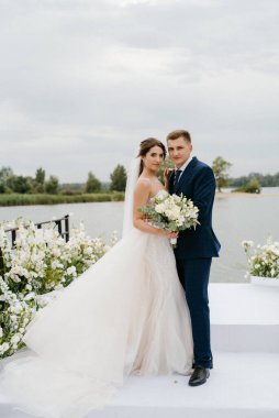 Gelin ve damat, nehrin kıyısındaki bir iskelede sarı bir günbatımının arkaplanına karşı.