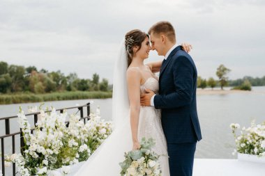 Gelin ve damat, nehrin kıyısındaki bir iskelede sarı bir günbatımının arkaplanına karşı.