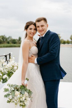 Gelin ve damat, nehrin kıyısındaki bir iskelede sarı bir günbatımının arkaplanına karşı.