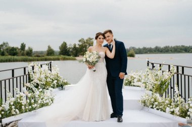 Gelin ve damat, nehrin kıyısındaki bir iskelede sarı bir günbatımının arkaplanına karşı.