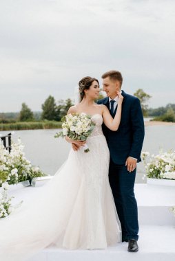 Gelin ve damat, nehrin kıyısındaki bir iskelede sarı bir günbatımının arkaplanına karşı.