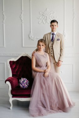 Bej takım elbiseli damat ve lavantalı gelin parlak stüdyoda dar bir gelinlik.